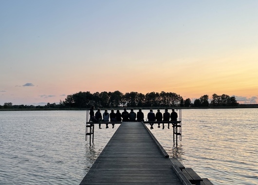 Ein Steg zieht geradeaus auf einen See. Am Ende des Stegs ist die Silhouette mehrerer sitzender Personen zu sehen. Im Hintergrund sieht man den Sonnenuntergang und eine Insel.