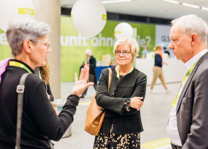 Man sieht zwei (ehemalige) Vorstandsmitglieder der GLS Bank auf einer Veranstaltung mit einer Mitarbeitenden reden.