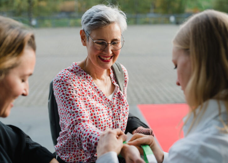 Man sieht drei Mitarbeitende der GLS Bank auf einer Veranstaltung. Einer Frau, die lächelt, wird ein Bändchen um das Handgelenk gelegt.