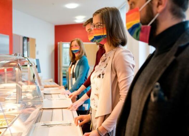 Man sieht Mitarbeitende der GLS Bank in der Kantine mit regenbogenfarbenen Masken Schlange stehen.