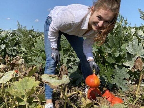 gls-bank-2022-nachhaltigkeitsbericht-biosphäre Eine Mitarbeitende der GLS Bank steht in einem Feld vor einer Kürbispflanze. Sie hält gebückt einen Kürbis in der Hand und lächelt in die Kamera.