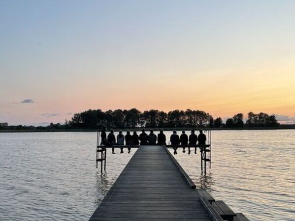 gls-bank-2022-nachhaltigkeitsbericht-wasser Ein Steg zieht geradeaus auf einen See. Am Ende des Stegs ist die Silhouette mehrerer sitzender Personen zu sehen. Im Hintergrund sieht man den Sonnenuntergang und eine Insel.