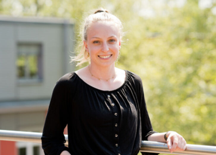 Sophia Orbach, die Leiterin des Projektes Nachhaltigkeitsbericht, blickt in die Kamera und lächelt. Im Hintergrund ist ein Teil des GLS Bank Gebäudes zu sehen und ein Baum.