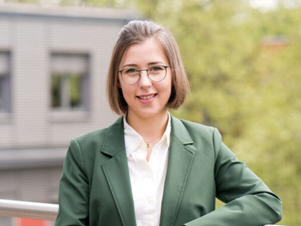 Aida Jukas, die Leiterin des Projektes Nachhaltigkeitsbericht, blickt in die Kamera und lächelt. Im Hintergrund ist ein Teil des GLS Bank Gebäudes zu sehen und ein Baum.