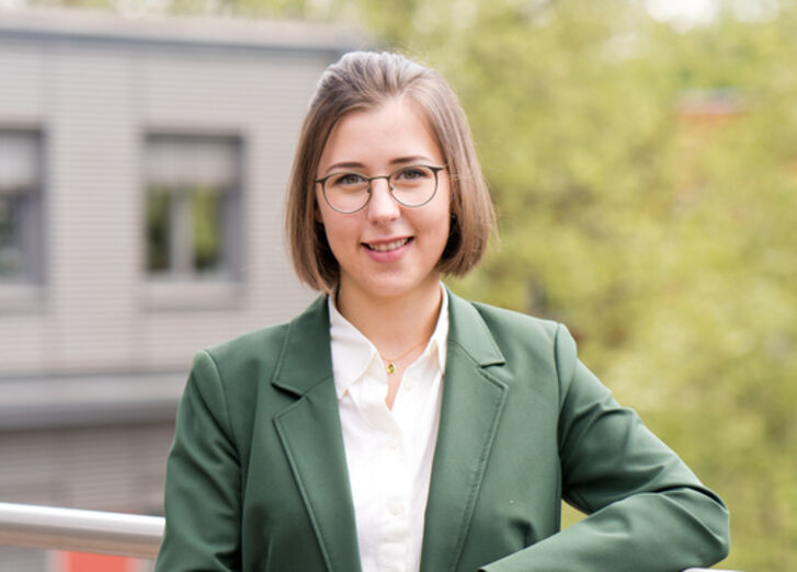 Aida Jukas, die Leiterin des Projektes Nachhaltigkeitsbericht, blickt in die Kamera und lächelt. Im Hintergrund ist ein Teil des GLS Bank Gebäudes zu sehen und ein Baum.